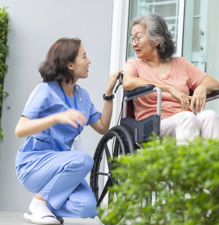 a caregiver kneeling to talk with a woman in a wheelchair outside a building